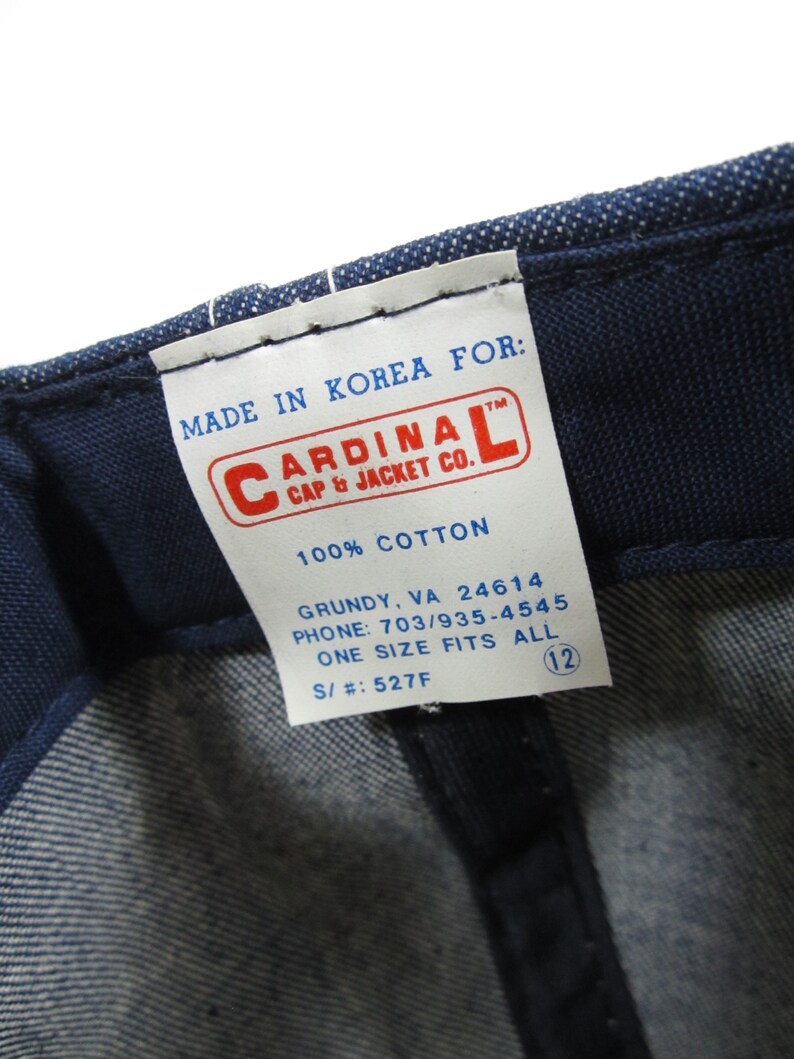 Puede incluir: Una etiqueta blanca con texto azul que dice "MADE IN KOREA FOR: CARDINAL CAP & JACKET CO. 100% COTTON GRUNDY, VA 24614 PHONE: 703/935-4545 ONE SIZE FITS ALL S/#: 527F" con un c&iacute;rculo azul que contiene el n&uacute;mero 12.