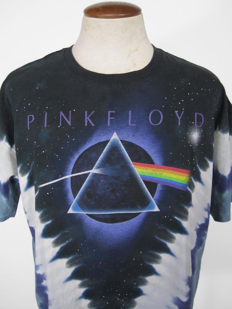 K&ouml;nnte beinhalten: Schwarzes T-Shirt mit Batikmuster und dem Pink Floyd Albumcover. Das Design zeigt ein schwarzes Dreieck, einen Regenbogen und den Namen der Band in lila Schrift. Das T-Shirt hat einen Rundhalsausschnitt und kurze &Auml;rmel.