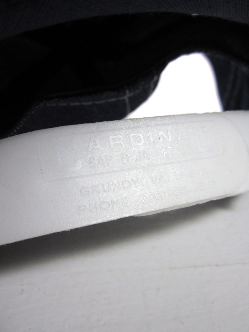 Puede incluir: Primer plano de una etiqueta blanca con el texto "ARDINA CAP & JACKET" y "GRUNDY, VA PHONE 703/937-****".