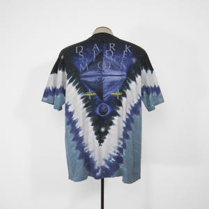 K&ouml;nnte beinhalten: Ein Batik-T-Shirt mit einem grafischen Design mit den Worten "DARK SIDE OF THE MOON" und einer Pyramide mit einem Auge. Das Shirt hat ein blaues, wei&szlig;es und schwarzes Farbschema.