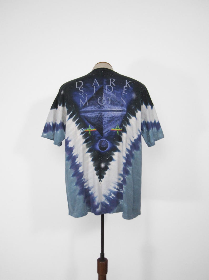 K&ouml;nnte beinhalten: Ein Batik-T-Shirt mit einem grafischen Design mit den Worten "DARK SIDE OF THE MOON" und einer Pyramide mit einem Auge. Das Shirt hat ein blaues, wei&szlig;es und schwarzes Farbschema.