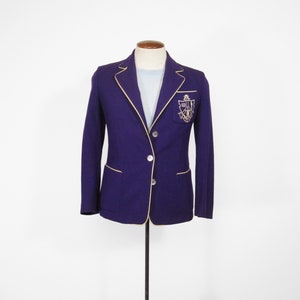 Puede incluir: Una chaqueta de color morado con ribetes dorados y un escudo bordado en oro en el lado derecho del pecho.