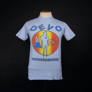 May include: A light blue short-sleeve t-shirt featuring a graphic print with the text DEVO and DUTY NOW FOR THE FUTURE. The design includes a central stylized figure, a laboratory flask with blue liquid, and an atom symbol inside a circular frame.