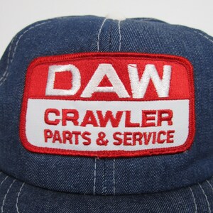 Puede incluir: Un gorro de camionero de mezclilla azul con un parche rojo y blanco que dice "DAW CRAWLER PARTS & SERVICE".
