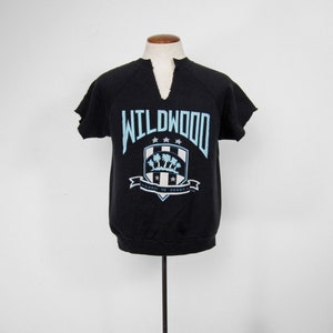 May include: Black short-sleeved sweatshirt with a blue and white graphic of a crest with palm trees and the text "WILDWOOD" and "COAST TO COAST".