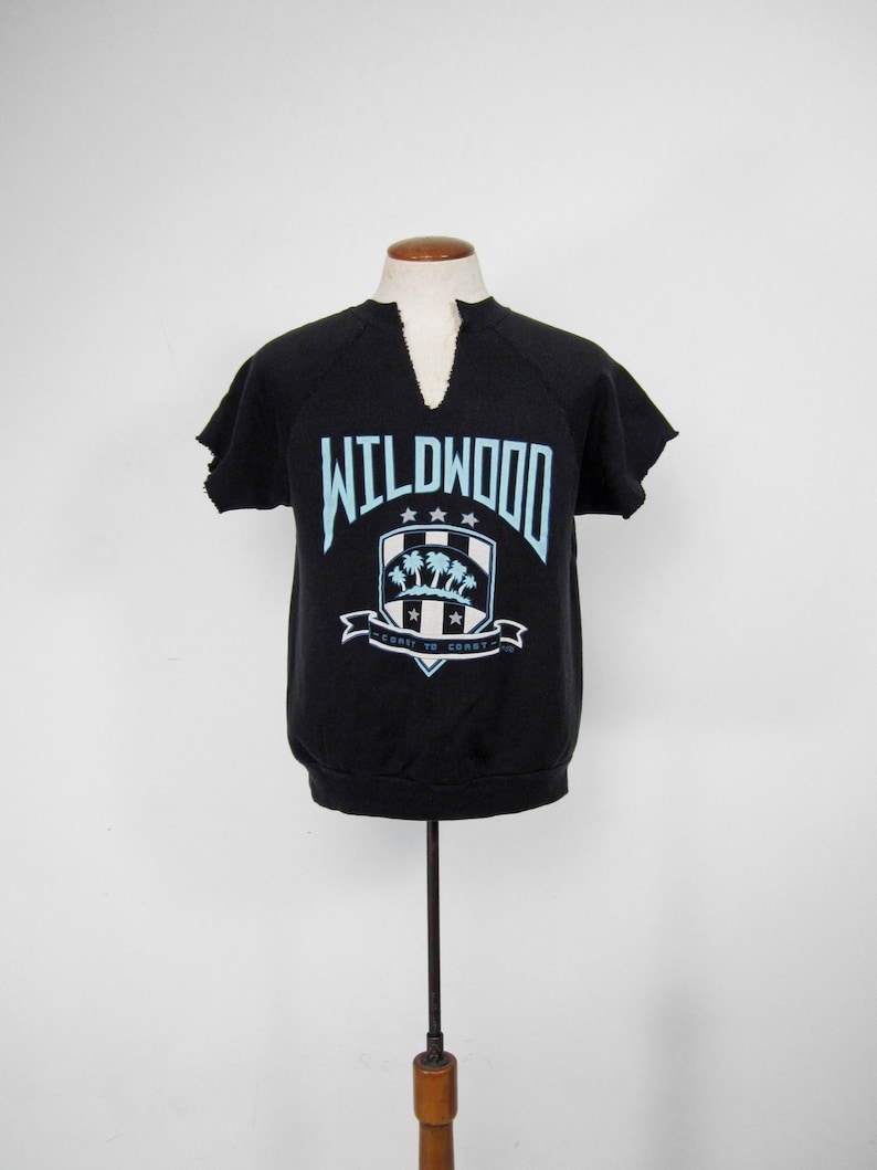 May include: Black short-sleeved sweatshirt with a blue and white graphic of a crest with palm trees and the text "WILDWOOD" and "COAST TO COAST".