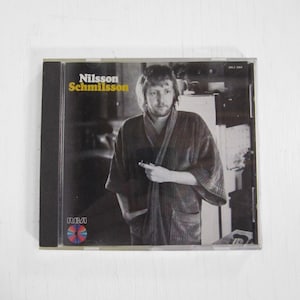 May include: A black and white photograph of a man wearing a dressing gown on a CD case. The album title is "Nilsson Schmilsson" and the record label is RCA.