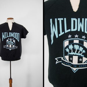 May include: Black short-sleeved sweatshirt with a blue and white graphic of a shield with palm trees and the text "WILDWOOD - COAST TO COAST -"