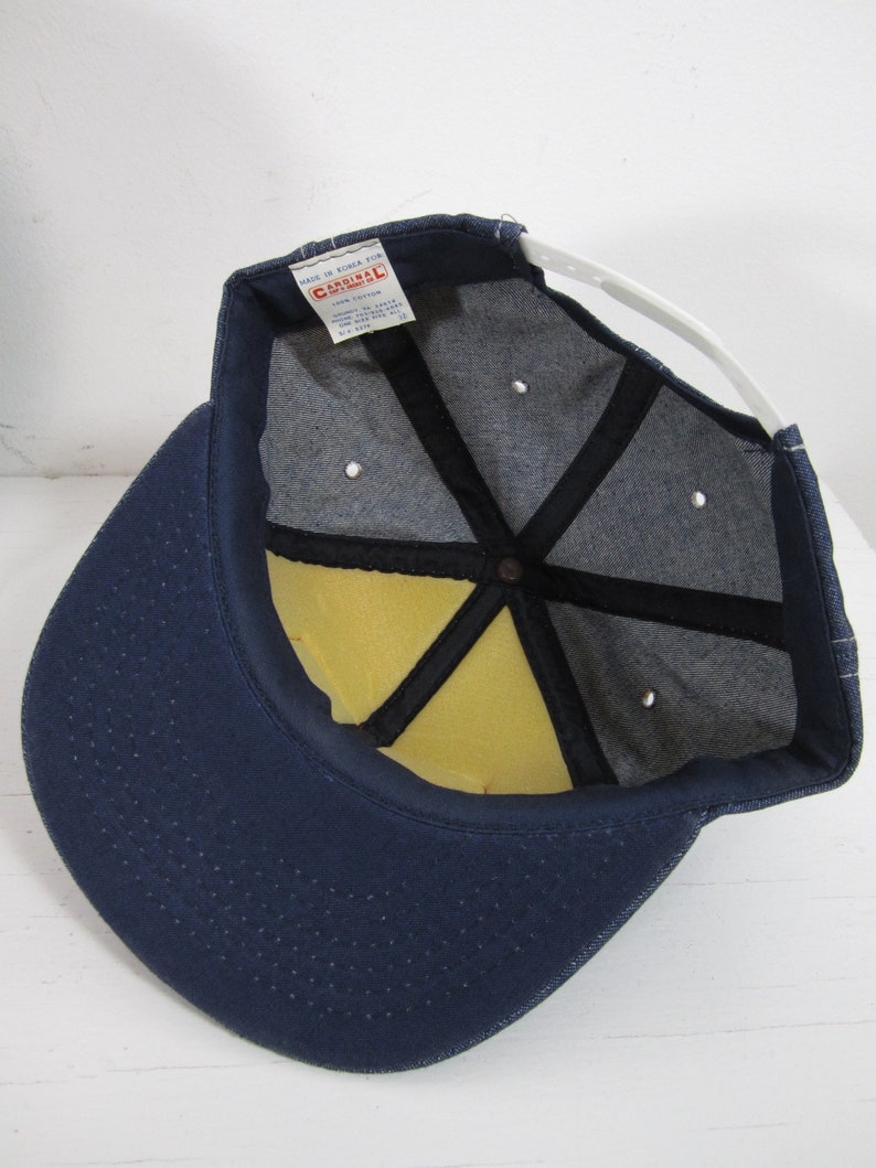 Puede incluir: Una gorra de b&eacute;isbol de mezclilla azul con una correa ajustable blanca y un interior de espuma amarilla. La gorra tiene una etiqueta que dice "Cardinal" y "Made in Korea for Cardinal Sportswear Co."