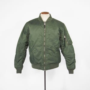 Könnte beinhalten: Olivgrüne Bomberjacke mit durchgehendem Reißverschluss, gerippten Bündchen und Saum. Die Jacke hat eine kleine Tasche am linken Ärmel. Präsentiert auf einer Schaufensterpuppe vor weißem Hintergrund.