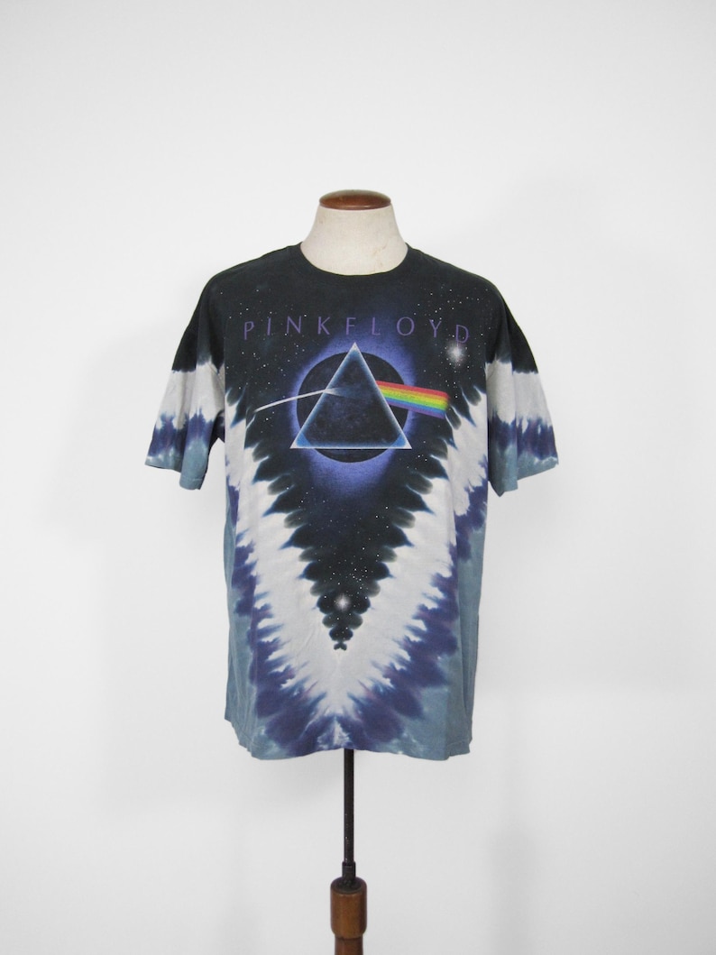 K&ouml;nnte beinhalten: Ein schwarzes T-Shirt mit Batikmuster und dem Pink Floyd Albumcover. Das Design zeigt einen schwarzen Hintergrund mit einem Dreieck, das einen Regenbogen bricht. Der Bandname PINK FLOYD ist oben in Wei&szlig; aufgedruckt.