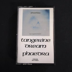 May include: A cassette tape with a white label featuring the album title "Phaedra" by Tangerine Dream. The label has a blue and white abstract design and the Virgin Records logo.
