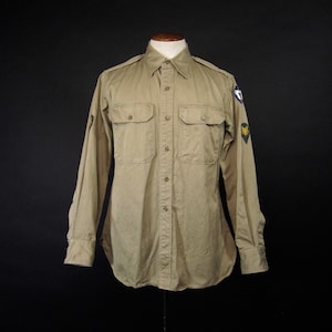 May include: A beige long-sleeved shirt with two patches on the left sleeve. The patches are green, yellow, and blue. The shirt has a button-down collar and a button-up front.