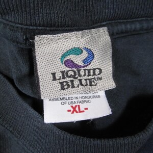 K&ouml;nnte beinhalten: Nahaufnahme eines schwarzen T-Shirt-Kragens mit Etikett. Das Etikett zeigt das "LIQUID BLUE"-Logo in T&uuml;rkis, Lila und Blau. Unter dem Logo steht "ASSEMBLED IN HONDURAS OF USA FABRIC" und "-XL-".