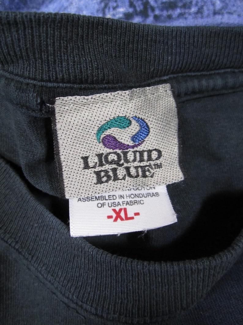 K&ouml;nnte beinhalten: Nahaufnahme eines schwarzen T-Shirt-Kragens mit Etikett. Das Etikett zeigt das "LIQUID BLUE"-Logo in T&uuml;rkis, Lila und Blau. Unter dem Logo steht "ASSEMBLED IN HONDURAS OF USA FABRIC" und "-XL-".