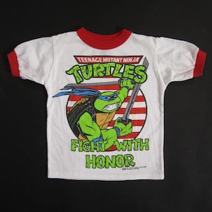 May include: White t-shirt with red trim featuring a Teenage Mutant Ninja Turtle graphic. The design includes the words "Teenage Mutant Ninja Turtles" and "Fight With Honor". The shirt has short sleeves.