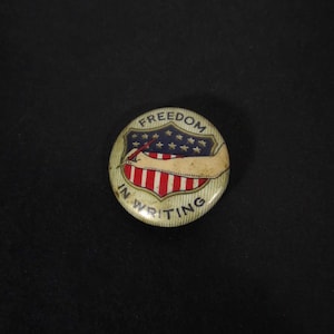 May include: A round button with a shield design featuring red and white stripes, blue stars, and the words "FREEDOM IN WRITING." The button has a vintage aesthetic.