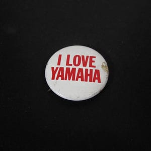 May include: A white, circular button with the red text "I LOVE YAMAHA". The button has a pin on the back and is slightly worn. The button is on a black background.