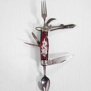 Puede incluir: Una multiherramienta vintage con un mango rojo con un diseño de dragón blanco. La herramienta incluye un tenedor, cuchara, cuchillo, abrelatas y otros implementos. La herramienta está abierta, mostrando todos sus componentes sobre un fondo blanco.