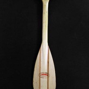 May include: A vintage wooden paddle with a light-colored finish and vertical wood grain. The paddle features a rounded blade with a label that reads "FEATHER BRAND". The handle has a hole for hanging.