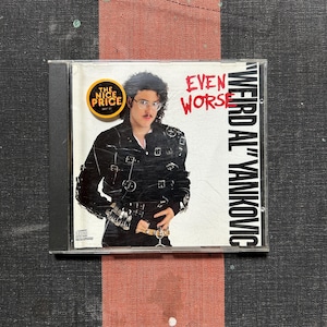 May include: A compact disc case featuring the album Even Worse by Weird Al Yankovic. The cover shows a person with curly hair and glasses wearing a black jacket with multiple buckles. A yellow circular sticker reads The Nice Price.