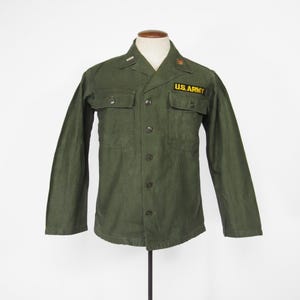 May include: Olive green military shirt with long sleeves, two chest pockets, and a button-up front. Features a "U.S. ARMY" patch and a small emblem above the right pocket. The shirt is displayed on a mannequin.