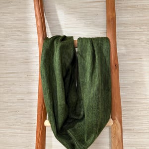 May include: A dark green infinity scarf draped over a rustic wooden ladder. The scarf has a soft, textured appearance and is made of a thick material. The ladder is made of natural wood and has a simple design. The background is a light-colored wall.
