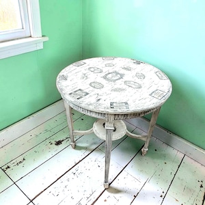 May include: A small, round, antique side table with a distressed white finish. The tabletop features a collage of vintage-style labels and designs. The table has a decorative apron and three curved legs, adding to its vintage charm. The table is approximately 76 cm high.
