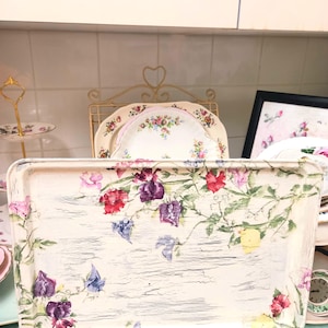 May include: Rectangular serving tray with a distressed white finish, decorated with a floral design featuring red, purple, and blue flowers. The tray has a vintage aesthetic, ideal for serving or display.