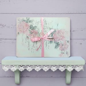 May include: A rectangular board with a light mint green background featuring a distressed finish and pink rose illustrations. A pink ribbon is tied around the centre of the board. The board rests on a light green shelf with white lace trim.