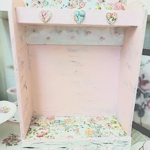 May include: A distressed pink wooden shelf with a floral patterned backing and base. The shelf has three heart-shaped embellishments and six small pegs. The interior is painted pink, and the shelf is designed for display.
