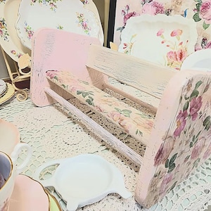 May include: A distressed pink wooden bench with a floral patterned seat and back. The bench is surrounded by vintage floral plates and teacups, creating a shabby chic aesthetic. The bench is approximately 30 cm long.
