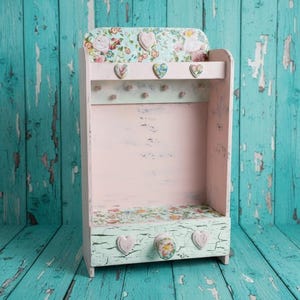 May include: A wooden wall-mounted organiser featuring a pale pink interior and a light green drawer with a cracked paint finish. The top shelf and drawer front are decorated with floral patterns and small heart-shaped accents and knobs.