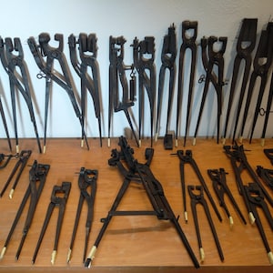 Op de afbeelding: Een verzameling antieke smidgereedschappen, waaronder tangen en andere gespecialiseerde instrumenten. De gereedschappen zijn gemaakt van donker metaal en zijn gerangschikt op een houten oppervlak. Sommige gereedschappen hebben lichtgekleurde tape aan de uiteinden.