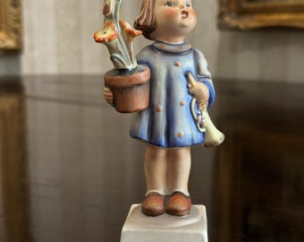 May include: A vintage porcelain figurine of a young child in a blue dress, holding a flower pot with a bird perched on top. The figurine has brown shoes and is standing on a white base. The child is looking upwards.