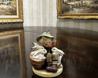 May include: A ceramic figurine of a young person in a green hat and brown boots, surrounded by two white rabbits and a basket of rabbits. The figurine is on a round, beige base. The background includes framed paintings.