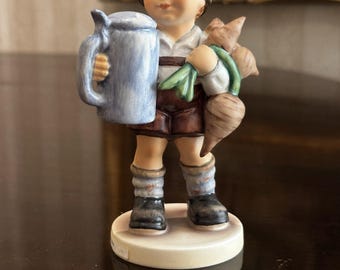 May include: A ceramic figurine of a young person in traditional Bavarian attire, holding a blue stein and a bundle of root vegetables. The figurine has a detailed painted face, a green hat, and brown shorts. The base is white.