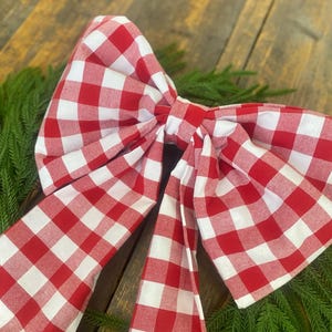 May include: A large, decorative bow made of red and white checkered fabric. The bow is set against a backdrop of green foliage and a wooden surface, creating a rustic and festive aesthetic. The bow's design is classic and suitable for various decorative purposes.