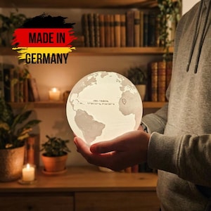 Könnte beinhalten: Eine leuchtende Globuslampe mit Weltkarten-Design wird in Händen gehalten. Die Lampe ist weiß und strahlt ein weiches Licht aus. Der Text "MADE IN GERMANY" ist zu sehen. Der Globus hat Koordinaten und eine Markierung.