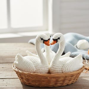 Puede incluir: Una cesta tejida contiene tres cisnes blancos de ganchillo. Los cisnes tienen picos naranjas y ojos negros, y están dispuestos en un abrazo amoroso. La cesta está sobre una superficie de madera, con una ventana y una manta azul al fondo.