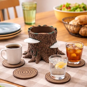 May include: A decorative tree stump centerpiece with white mushroom accents, surrounded by wooden coasters with concentric circle designs. A white mug of coffee, glasses of water and iced tea, and a bowl of salad are on a checkered tablecloth.