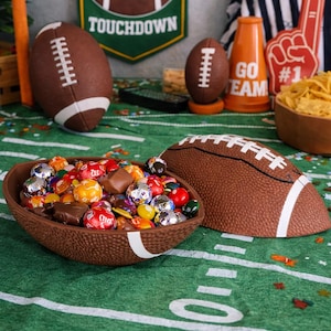 May include: A football-themed display with a brown football-shaped bowl filled with colorful candies. Other football decorations include a banner that says "Touchdown", a foam finger, and a wooden bowl of chips. The scene is set on a green field.