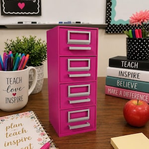 May include: A vertical desktop storage unit featuring four stacked drawers in a vibrant magenta color. Each drawer is equipped with a rectangular silver handle and a label holder. The unit has a smooth, solid finish and a rectangular frame.