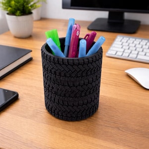 May include: A black pen holder shaped like a tire, filled with colorful pens and markers. The desk organizer is made of a textured material and sits on a wooden desk. A small plant and computer are in the background.