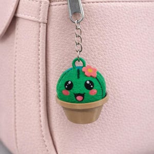May include: A green cactus keychain with a smiling face, pink cheeks, and a small pink flower on top. The cactus is in a brown pot and attached to a silver chain and zipper. The background is a pink purse.