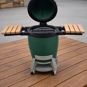 May include: A green ceramic grill with a black lid and side shelves. The grill sits on a gray stand with wheels and is open, revealing a metal grate. The grill is on a wooden table.