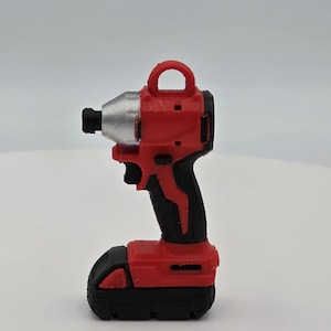 May include: A miniature red and black cordless impact driver replica featuring a silver-coloured nose cone, a black trigger, and a small loop at the top for hanging. The tool has a textured black handle and a black battery base, rendered in a solid form.