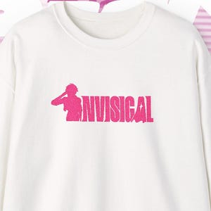 Puede incluir: Sudadera blanca de cuello redondo con el texto rosa "NVISIGAL" y un gráfico. El fondo presenta un diseño rosa y blanco con personajes de dibujos animados. La esquina inferior derecha tiene el texto "CAM 01".