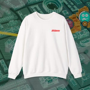 Peut inclure: Un sweat-shirt blanc à col rond et à manches longues. Les lettres rouges "SDN" sont imprimées sur le haut du côté gauche de la poitrine. Le fond est un motif de carte vert et turquoise.