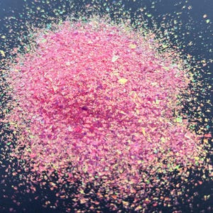 May include: A pile of fine, iridescent pink flakes with a reflective, metallic finish. The small, irregular shards are concentrated in the center and scatter outward, displaying subtle shifts of gold and green light against a dark background.